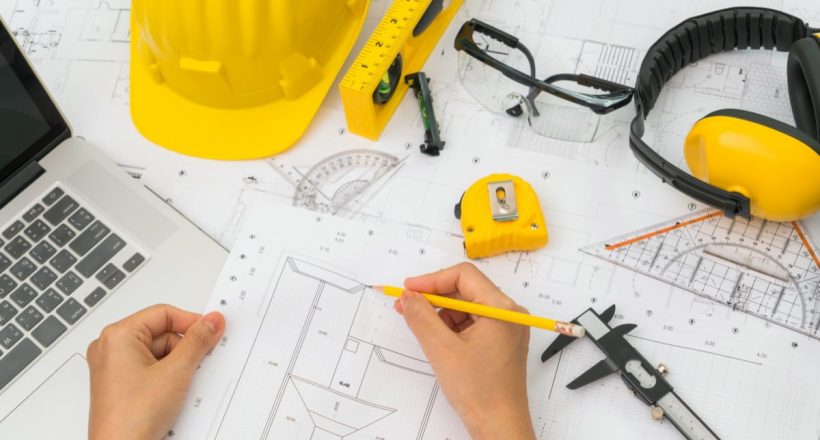 Hand over Construction plans with yellow helmet and drawing tools on blueprints